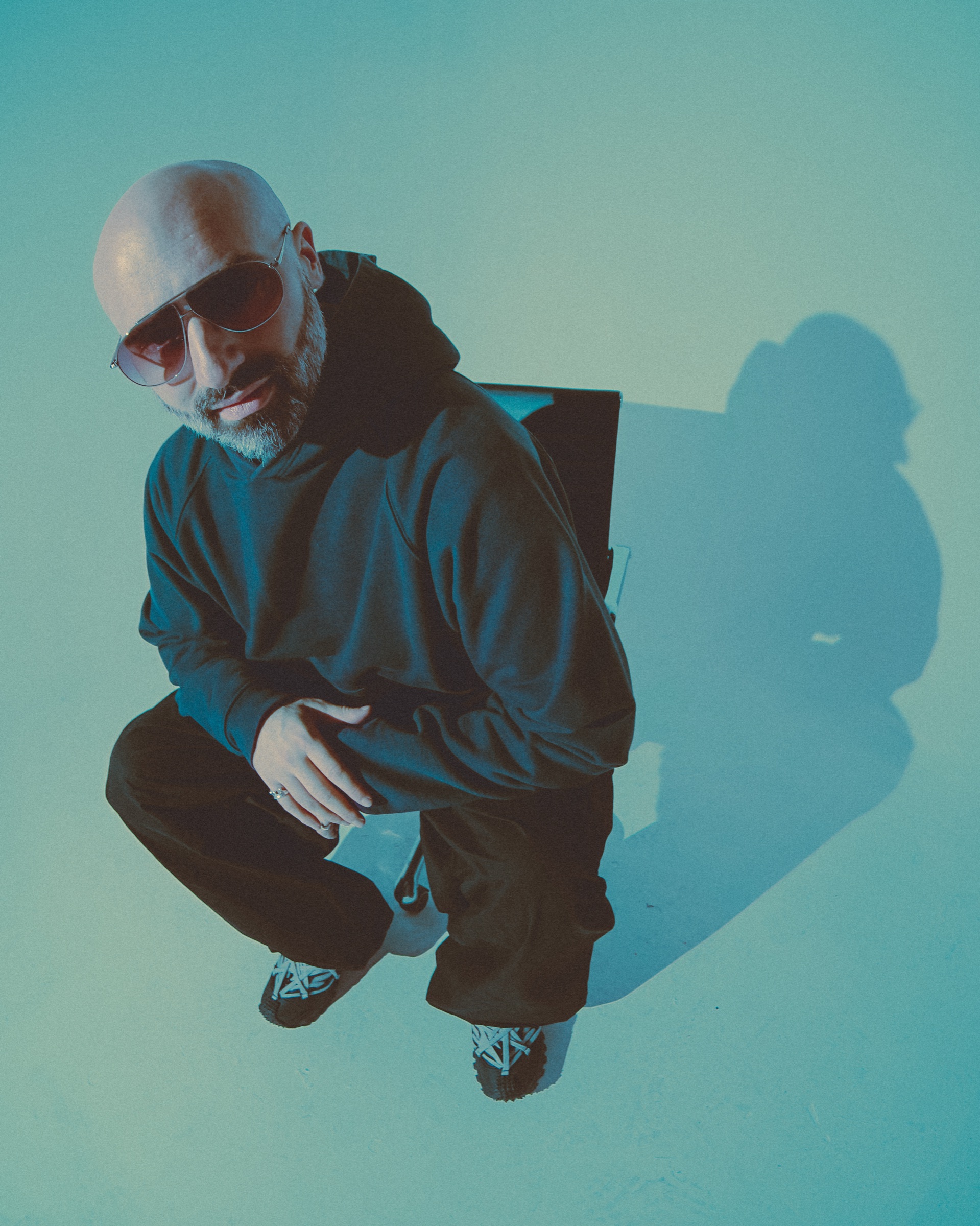 DECAP crouched against a teal wall in a dark jacket, sunglasses, his shadow beside him.